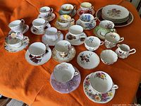 Set of 17 teacups and saucers of various fine china patterns displayed on an orange fabric surface.