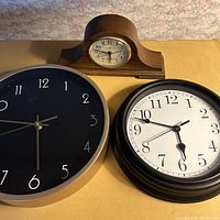 Three clocks displayed: wooden Howard Miller mantle clock centered on top, black Baitu wall clock with gold rim on left, black Vivatime wall clock with white face on right.