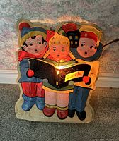 Front view of the light up Christmas carolers decoration showing three children in winter outfits holding a songbook that is lit from inside.