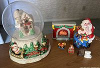 Snow globe featuring snow-covered house inside a dome, with a base decorated with greenery, Santa's sleigh, and presents. Also pictured are several holiday figurines including Santa, fireplace, toy horse, teddy chest, and kitten clock.