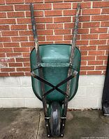 Full view of the green metal wheelbarrow showing the bucket, wooden handles, and front tire, leaning against brick wall.