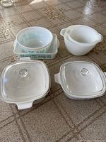 Seven white and patterned serving bowls arranged on patterned tablecloth, including two CorningWare square bowls with clear lids, one large white round bowl, and three Pyrex bowls with motifs.