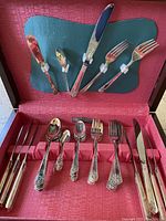 Open wooden box showing silver plated flatware with forks, spoons, knives arranged inside lined with red fabric. Handles have grape cluster pattern.