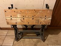 Front view of the collapsible workbench in folded position showing the wooden top with holes and metal frame.
