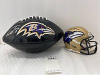 Front view showing black Ravens football with white laces and synthetic leather surface, beside gold mini helmet