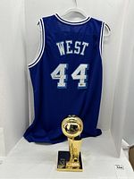 Full view back of blue Jerry West #44 jersey on hanger with gold-tone replica championship trophy in front