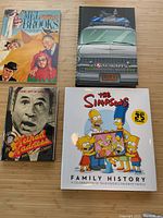 Four books open to show front covers: The Simpsons Family History hardcover, Ghostbusters cartoon hardcover, Mel Brooks spoof softcover, In Method Maddness softcover