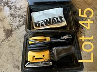 Open carrying case showing yellow DeWalt sander with attached dust bag and coiled power cord, along with a folded DeWalt manual inside.