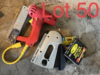 Image showing two staplers (a large red heavy-duty staple gun and a smaller silver hand stapler) along with two boxes of staples on a concrete surface.