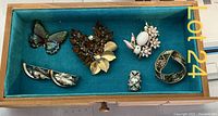 Six decorative brooches laid flat in a teal fabric-lined box, displaying different shapes including butterfly, bird, floral cluster, abstract, and ring designs.