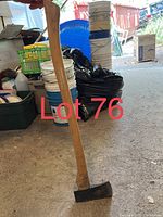 Photo showing an ax with a wooden handle standing upright indoors on concrete floor near storage items.