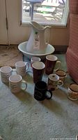 Photo showing the white ceramic wash basin and matching pitcher with 10 assorted mugs in front in varied designs and sizes on carpet