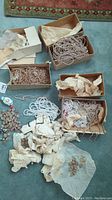 Wide shot showing several cardboard boxes containing multiple strings of faux pearl beaded necklaces wrapped in tissue paper with some scattered pieces of costume jewelry on carpet.