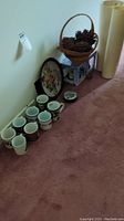 Photo showing a set of eight Christmas themed mugs arranged on pink carpet next to a basket of pine cones and other holiday decor