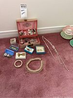 Full view of jewelry box open with clip-on earrings, brooches, pins inside and various necklaces, bracelets, and boxes around it on carpet.