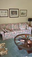 Front view of a three-seat fabric couch with pastel color pattern, four matching throw pillows, and fabric arm covers, set against a wall with three framed artworks.