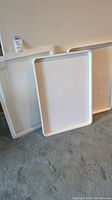 White rectangular food service trays leaning against wall, showing front surfaces and edges.
