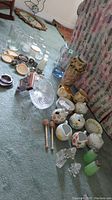 Photo showing assorted salt and pepper shakers, ceramic sugar jars and creamers, plates, and large glass pitcher on carpet near couch and curtain.