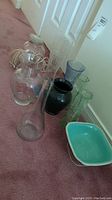 Multiple glass vases of different shapes and sizes including clear, black, green, and blue glass, and an oil lamp chimney on the carpet