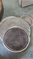 Photo showing multiple metal pizza pans stacked, all with perforated mesh design and raised edges, displaying signs of use and need for cleaning.