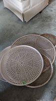 Photo showing several round perforated aluminum pizza pans stacked, varying sizes visible, some with handles.