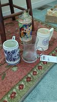 Four different steins on carpet: one thin clear glass with etched boat, one colorful ceramic with metal lid (Niagara on The Lake), one hand-painted Holland ceramic with metal lid, one white music box stein with handle and blue decal.
