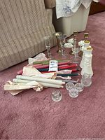 Overview of assortment of taper candles, glass and metal candlesticks, small glass ashtrays, and white ceramic candle holders.