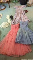 Three vintage dresses laid out on a floor: a long pink lace-trimmed dress, a coral strapless tulle dress, a light blue strapless dress, with white crocheted hat and gloves