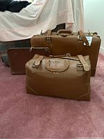 Three pieces of vintage brown luggage on carpet: 27-inch suitcase with broken strap, duffle bag, leather folio bag