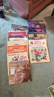 Photo of arranged vintage vinyl soundtrack records on floor with a couch and chair visible. Shows colorful record covers of titles including Pinocchio, Oklahoma, The Sound of Music, Carousel, Mary Poppins, Doctor Dolittle, The King and I.