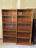Two tall particle board bookshelves side by side against a wall with five shelves each and a medium brown wood grain finish.
