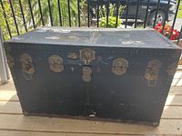 Front view of the closed antique trunk showing black exterior with metal hardware and latches, areas of wear and damage visible.