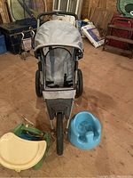 Photo showing the gray jogging stroller with three wheels, the blue Bumbo chair, and the green travel high chair with yellow tray on a wooden floor in a storage or basement area.