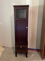 Tall dark wooden bathroom cabinet with glass pane door and lower drawer, shown from front.
