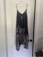 Navy blue romper hanging on a white hanger against a white door. Romper has white and beige paisley and floral patterns at the bottom, thin spaghetti straps, and a curved neckline. Tags visible on the front strap indicating it is new and unworn.