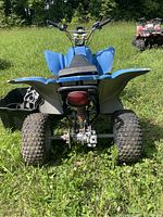 Rear view of blue children's four-wheeler with black seat and large knobby tires, engine area showing disassembly, black bucket of engine parts near rear left tire.