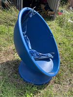 Full view of blue plastic egg chair on grass with torn blue canopy inside seat area.