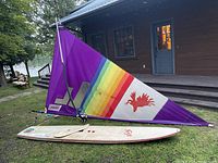 Side view of the windsurfing sail and board set up on the ground next to a house. The sail is purple with a rainbow stripe and a red phoenix emblem.