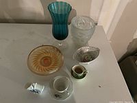 Photo showing six assorted glass and porcelain pieces including a teal blue vase, frosted floral glass, amber glass bowl, floral porcelain dishes, and small creamer