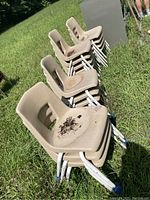 Stacked beige hard plastic chairs with metal legs on grass, showing dirt and rust spots.