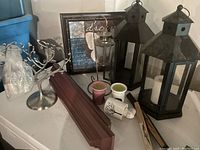 Photo shows two large black metal lanterns, framed wall art, metal jewelry tree, small candle holders, clear glass oil lamp, wooden wall shelf, and two decorative fans.