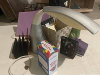 Silver flexible-neck desk lamp, black and purple wire mesh organizers, woven basket with sticky notes, slim jewel CD cases, purple folder, and CDs visible on a white surface.
