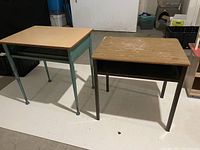 Photo of two vintage single-student school desks showing wooden tops and metal frames. Left desk has green frame, right desk has dark brown frame and visible surface wear.