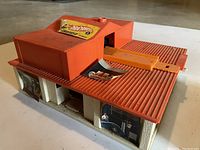 Front view of vintage orange plastic Hot Wheels play garage with worn and peeling original logo stickers on roof and side.