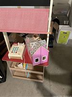 Wooden dollhouse with pink walls and gray roof displayed alongside the toy cash register and a portion of a wooden shelf unit nearby