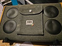 Front view of Marley Bluetooth speaker showing fabric exterior, four speaker grilles, and flap with Marley logo.