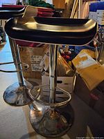 Photo showing two black seat bar stools with chrome bases and footrests in a storage area with various boxes and items around them.