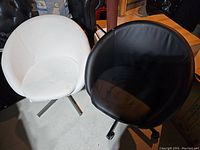 Two round chairs, one black leather with wheels and one white fabric missing wheels, on a concrete floor in storage area.