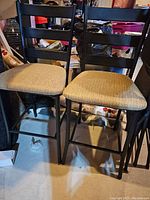 Two black wooden chairs with slatted backs and beige upholstered seats, placed on concrete floor surrounded by various items.