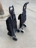 Two black fabric shopping buggies with metal frame and wheels, viewed from the side leaning against each other.